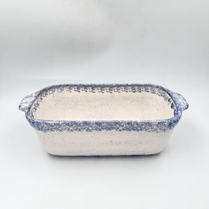 May include: A rectangular, off-white ceramic baking dish with a speckled surface and blue floral trim. The dish has two small handles and is suitable for baking casseroles and other dishes. The overall design is classic and rustic.