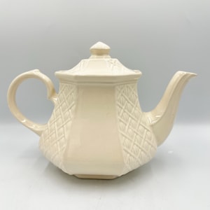 May include: Cream-colored ceramic teapot with a lid and a curved handle. The teapot has a textured diamond pattern on the body and a spout for pouring. The teapot is designed for brewing and serving tea.