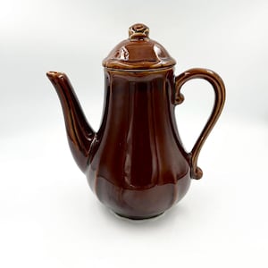 May include: A glossy, dark brown ceramic teapot featuring a vertical ribbed body, a curved handle, a long tapered spout, and a matching lid with a small knob handle on top. The teapot has a smooth, reflective finish and a rounded base shape.