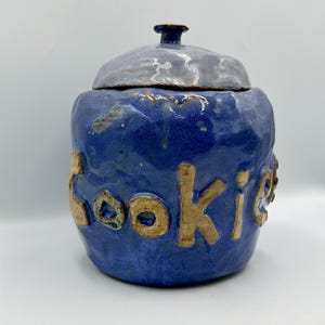 May include: A handmade ceramic cookie jar with a blue base and a gray lid. The word "cookie" is written in raised, gold-colored letters on the jar's front. The jar has a small, dark brown knob on top.