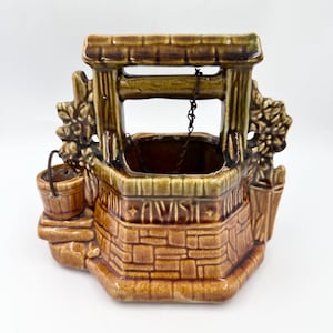 May include: A glazed ceramic wishing well planter in shades of brown and tan. The structure features a stone-textured base, a roof supported by two posts, a small attached bucket, and a thin metal chain hanging from the center of the roof.