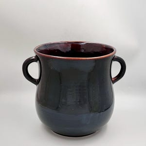 May include: A dark, glazed ceramic vase with two handles. The exterior is a deep, glossy black, while the interior and rim have a rich, burgundy hue. The vase has a rounded body and a wide opening.