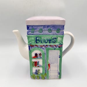 May include: A ceramic teapot shaped like a shop, featuring the text "Boots" in green. The teapot has a white spout and handle, with a green and blue facade, and a light purple lid. The shop design includes shelves with decorative items.