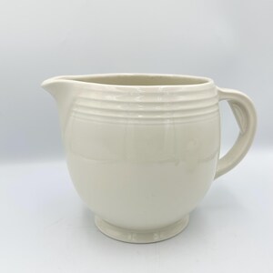 May include: Cream-colored ceramic pitcher with a rounded body, a spout, and a curved handle. The pitcher has horizontal ridged detailing around the top. The base is a small, circular pedestal.