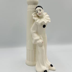 May include: A cream-colored ceramic figurine of a Pierrot clown leaning against a column. The clown is dressed in a white outfit with a ruffled collar, black accents, and a black cap. The figurine is a decorative item.