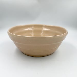 May include: A light beige ceramic bowl with a smooth, glossy finish and a wide, rounded rim. The exterior of the bowl features three horizontal, recessed grooves circling the body, providing a subtle texture to the otherwise simple, circular design.