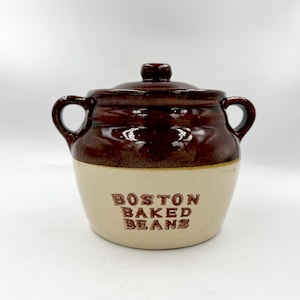 May include: A two-handled ceramic bean pot with a lid, featuring a glossy dark brown glaze on the upper portion and a cream-colored base. The text BOSTON BAKED BEANS is embossed in brown lettering on the front of the pot, which has two side handles.