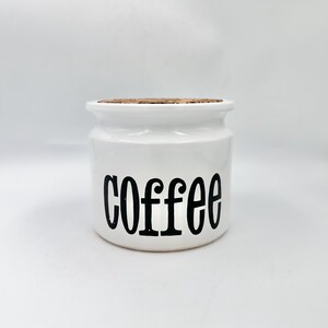 May include: White ceramic coffee canister with a cork lid. The word "coffee" is printed in black, bold letters on the front of the canister. The canister is cylindrical with a slightly flared top.