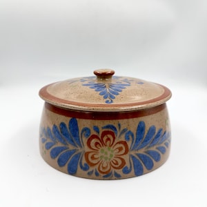 May include: A ceramic lidded container with a tan base and lid, adorned with blue floral patterns and a central flower design in orange, green, and brown. The rim and knob are brown.