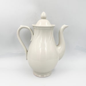 May include: Cream-colored ceramic teapot with a lid and a curved handle. The teapot has a bulbous body and a long, curved spout. The lid is topped with a small, decorative finial. The teapot is designed with a swirl pattern.