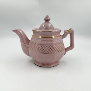 May include: A light pink ceramic teapot featuring a hexagonal body, a matching lid with a finial, and a curved handle. The teapot is decorated with gold-colored trim along the rim, spout, and handle, and a gold lattice pattern around the center.