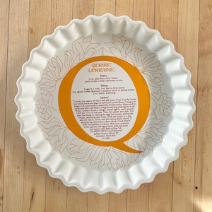 May include: White ceramic quiche dish with a scalloped edge and floral design. The center features a recipe for Quiche Lorraine in orange and black text. The dish is on a wooden surface.