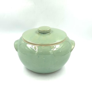 May include: A round, light sage green ceramic pot with a matching lid and two side handles. The surface has a smooth, glossy finish with subtle tonal variations in the glaze, creating a uniform appearance across the body and the circular lid.