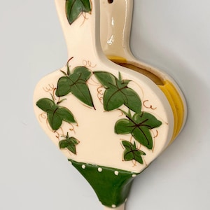 May include: A vintage ceramic wall pocket vase with a cream-colored base and green ivy leaf design. The vase has a green and white polka-dot trim and a yellow interior. It is designed to hold flowers or other decorative items.