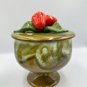 May include: A ceramic jar with a lid, featuring a green and brown glaze. The lid is adorned with three red, glossy ceramic strawberries and green leaves. The jar has a pedestal base and a rounded body.