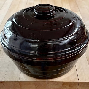 May include: A dark brown, glazed ceramic casserole dish with a matching lid. The dish has a round shape with horizontal ridges and a glossy finish. The lid features a small, circular handle. The dish sits on a light-colored wooden surface.