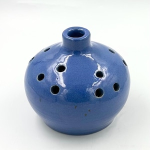May include: A round, blue ceramic flower frog with a glossy finish. The object features a central opening at the top and multiple smaller, circular holes distributed across its upper surface, designed for arranging individual flower stems.