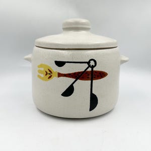 May include: A cream-colored ceramic lidded pot with a speckled finish and two side handles. The front features a graphic illustration of a yellow fork, a brown textured utensil, and three black measuring spoons arranged in a circular pattern.
