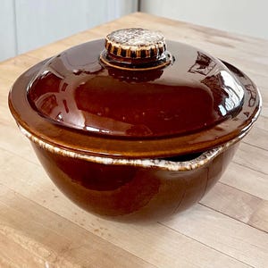 May include: A brown ceramic casserole dish with a lid. The dish has a glossy finish and a white rim. The lid features a decorative handle. The dish is sitting on a wooden surface.