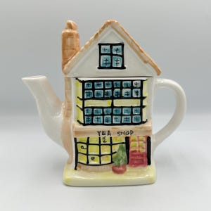 Peut inclure: Théière en céramique en forme de salon de thé. Elle présente un toit blanc, des murs jaunes et blancs, et une cheminée. L'inscription "TEA SHOP" est imprimée sur le devant. Bec et anse blancs.