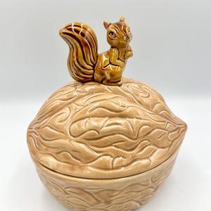 May include: A ceramic nut-shaped container with a squirrel figurine on top. The container is light brown with a textured surface, and the squirrel is brown with a bushy tail. The lid fits snugly, creating a decorative and functional piece.