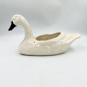 May include: A ceramic swan-shaped planter featuring a cream-colored body with a distinct dark crackle glaze finish. The swan has a black beak and a hollowed-out center for holding items, with a long, curved neck and detailed wing textures.