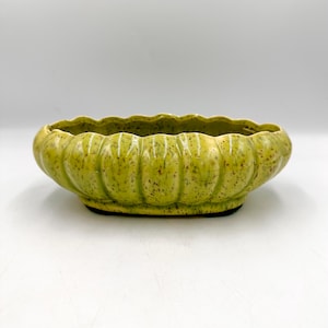 May include: A vintage, oval-shaped ceramic bowl in a speckled, light green glaze. The bowl has a scalloped rim and a dark base. The exterior features vertical ridges, adding texture to the piece. This decorative bowl is suitable for various uses.