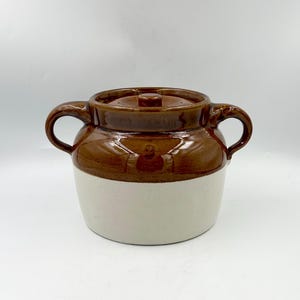 Op de afbeelding: Een tweekleurige keramische pot met deksel, voorzien van een glanzend bruin glazuur aan de bovenzijde en een matte, gebroken witte afwerking aan de onderzijde. Het vat heeft een brede, ronde vorm met twee gebogen handvatten en een kleine knop.