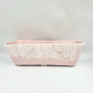 May include: A rectangular, pink ceramic planter with a textured, speckled design. The planter has a light pink base and a white, speckled upper section. The planter is designed for indoor or outdoor use.