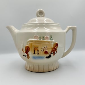 May include: Cream-colored ceramic teapot with a lid and handle. The teapot features a decorative scene with a cabin, chairs, and a fireplace. The design includes brown, blue, and pink accents. The teapot has a pedestal base.