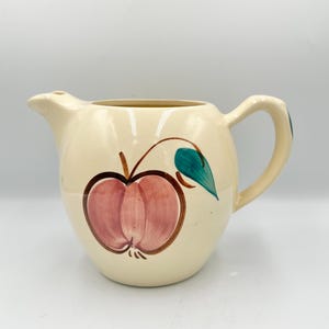 May include: Cream-colored ceramic pitcher with a round body, spout, and handle. The pitcher is decorated with a large pink apple design with a brown outline and a green leaf. A classic kitchenware item.