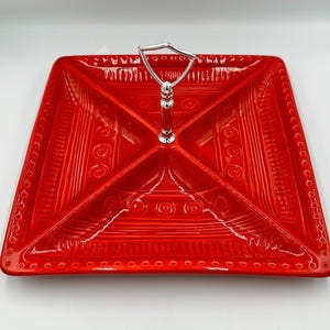 May include: A square, red ceramic serving dish with a silver handle. The dish is divided into four sections and features a textured design with circles and lines. The handle is a decorative element.