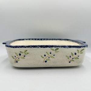 May include: Rectangular ceramic baking dish with a speckled cream-colored base and a dark blue rim. The dish features hand-painted green leaves and blue berry sprigs. It has two small handles on either side.