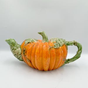 Puede incluir: Una tetera de cerámica con forma de calabaza. La tetera es de color naranja con detalles verdes en el asa, el pico y el tallo. La calabaza tiene estrías verticales y un acabado brillante. Un artículo decorativo para la cocina o el comedor.
