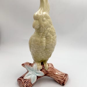 May include: A ceramic figurine of a parrot perched on a brown branch with a light blue flower. The parrot is a light green color with textured feathers and a crest. The base is brown with a light blue flower.