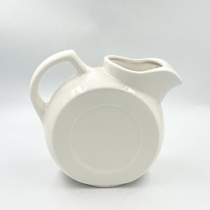 May include: A glossy off-white ceramic pitcher with a rounded, circular body, a curved handle on the left side, and a prominent pouring spout on the right. The surface is smooth and uniform in color, featuring a distinct circular indentation.