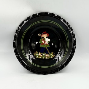 May include: A round black ceramic bowl with a scalloped edge and a central hand-painted illustration of a figure in a red hat and green jacket holding a wooden staff and a small white lamb, surrounded by green foliage and small white flowers.