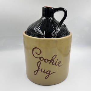 May include: A tan ceramic cookie jug with a black lid and handle. The jug has the words "Cookie Jug" in a brown script. The lid has a textured, leaf-like pattern.