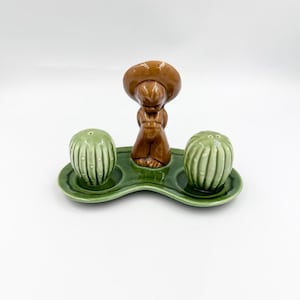 May include: A ceramic condiment set featuring two light green, ribbed cactus-shaped shakers and a central brown figure wearing a wide-brimmed hat. These pieces are arranged on a matching green, curved, kidney-shaped base with two circular indentations.