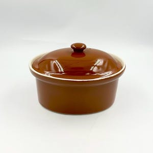 May include: A round, brown ceramic casserole dish with a matching lid. The dish features a smooth, glossy finish and a white interior rim. The lid has a small, centered knob handle, and the overall design is simple, functional, and cylindrical.