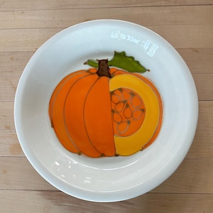 May include: A white ceramic plate with a hand-painted pumpkin design. The pumpkin is orange and yellow with a brown stem and green leaf. The plate is round and has a slightly raised edge.