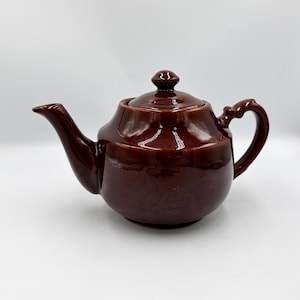 May include: A brown ceramic teapot with a lid, spout, and handle. The teapot has a glossy finish and a rounded body. The handle is curved, and the spout is slender. The lid has a small knob on top. The teapot is designed for brewing tea.