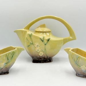 Puede incluir: Un juego de té de cerámica amarilla vintage con una tetera y dos azucareros/cremeros. La tetera tiene un asa curva y tapa. Diseños florales y de hojas decoran el juego. El juego tiene un esquema de color de dos tonos con una base más oscura.