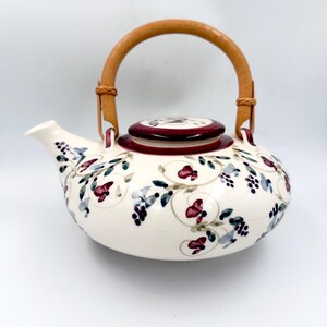 May include: A ceramic teapot with a cream-colored base featuring a hand-painted pattern of dark red flowers, blue leaves, and small black berries. The teapot has a matching lid with a red rim and a curved, light brown wooden swing handle.