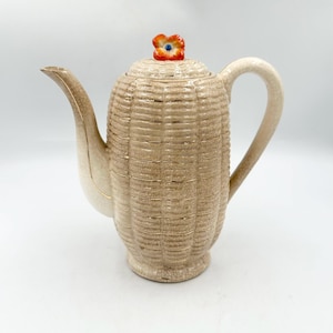 May include: A ceramic teapot with a beige, crackled glaze finish and a textured surface resembling corn kernels. The lid features a small, orange flower-shaped knob with a blue center. The teapot has a curved handle and a long, tapered spout.