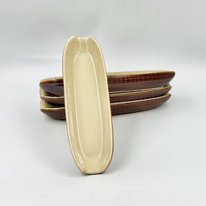 May include: A set of four elongated ceramic dishes with a smooth cream-colored interior and a textured, dark brown exterior. The dishes are oval-shaped with slightly raised edges, stacked horizontally with one dish standing upright in the front.
