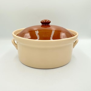 May include: A tan ceramic casserole dish with a brown lid and a small knob. The dish has two handles and a rounded shape. The lid is a glossy brown color.