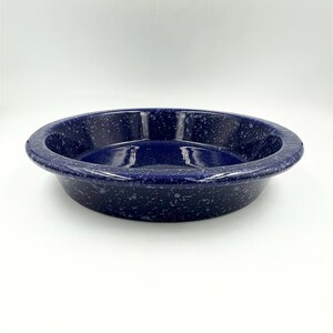 Puede incluir: Un molde para tarta redondo y profundo con un color base azul oscuro y un patrón moteado en blanco y azul claro en todas las superficies exteriores e interiores. El plato tiene un borde ancho, liso y redondeado, y un acabado brillante.