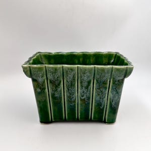 May include: A rectangular, green ceramic planter with a textured, ribbed design. The planter has a glossy finish and sits on four small feet. The dark green color and unique shape make it a decorative item for plants or other items.