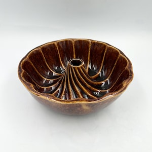 May include: A round, brown ceramic baking mold featuring a glossy, mottled glaze finish. The interior of the bowl has a distinct fluted, spiral design radiating from a central hollow tube, creating a decorative pattern for molded food items.